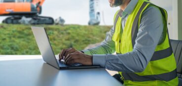 Person an einem Laptop, die Person trägt ein Hemd und eine Warnweste, im Hintergrund ist ein Hitachi Kettenbagger zu sehen